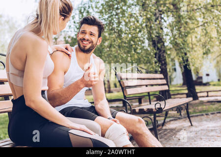 Feriti sportivo sorridente e mostrando un pollice alzato durante la seduta sul banco vicino a giovani sportive Foto Stock