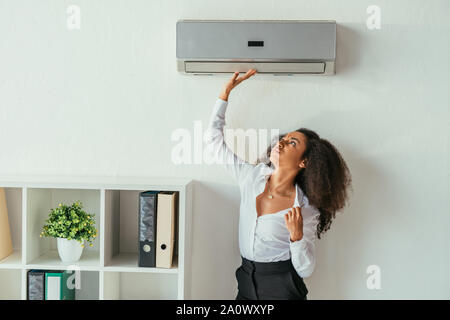 Piuttosto americano africano imprenditrice toccando il condizionatore d'aria mentre la sofferenza dal calore in office Foto Stock