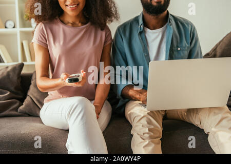 Vista parziale di African American uomo utilizzando computer portatile mentre è seduto sul lettino nelle vicinanze African American donna tenendo il condizionatore d'aria il dispositivo di controllo remoto Foto Stock