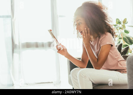Scioccato americano africano donna guardando il termometro mentre la sofferenza dal calore estivo a casa Foto Stock