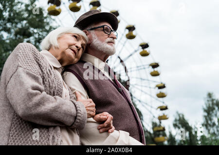 Basso angolo di visione della donna senior abbracciando il marito vicino a ruota panoramica nel parco di divertimenti Foto Stock