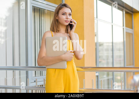 Affascinante giovane donna, studente in casual tuta gialla parlando al telefono cellulare e computer di contenimento in altra parte permanente ufficio all'aperto, college, Foto Stock