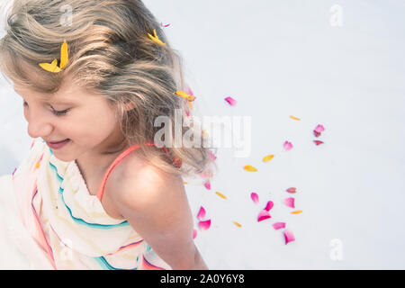 Close up ritratto di adorabili poco ragazza seduta con stilizzata lunghi capelli biondi decorato con fiori Foto Stock