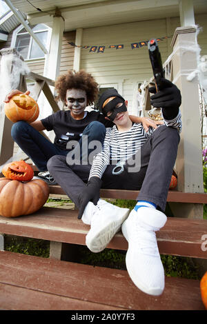 Angolo basso ritratto di due ragazzi adolescenti che indossano costumi di Halloween seduti sulla veranda della casa decorata e guardando la telecamera, spazio di copia