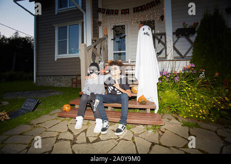 A piena lunghezza ritratto di due ragazzi che indossano costumi di Halloween in posa all'aperto seduti sulla veranda insieme nel buio, shot con flash, spazio di copia Foto Stock