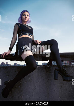 Ritratto di bella sorridente ragazza gotica contro il cielo al tramonto del tempo. Goth pastello con violetto (viola) capelli vestiti di nero Foto Stock