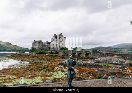 Dornie, Scozia; 27 Agosto 2019: a Scottish bagpiper giocando il suo strumento musicale al di sopra di Eilean Donan Castle Foto Stock