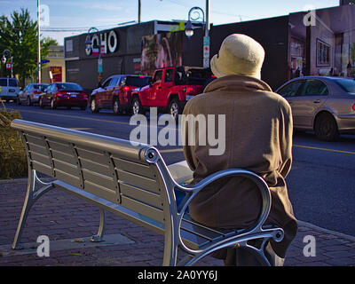 Signora avvolto in un abbigliamento caldo e un cappello di lana si siede su una panchina in attesa per il numero 85 bus, Preston Street, Ottawa, Ontario, Canada. Foto Stock