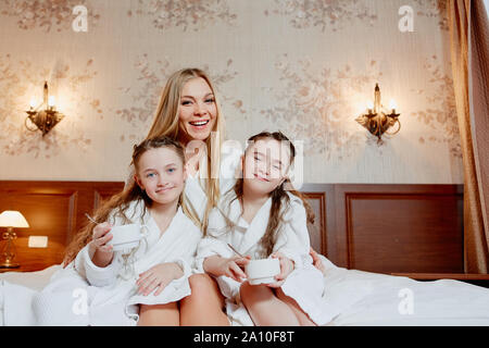 Bella e felice madre e le sue due figlie, abbracciare e ridere. Sedetevi e rilassatevi su un letto in camera accogliente. La prima colazione. Foto Stock