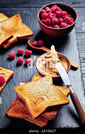 Close-up di aprire i sandwich di pane tostato con dolci burro di arachidi e lamponi freschi su un nero ardesia il vassoio su un legno nero tavolo da cucina, ver Foto Stock