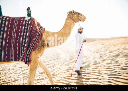 Uomo che indossa gli abiti tradizionali, tenendo un cammello sulla sabbia del deserto, a Dubai Foto Stock