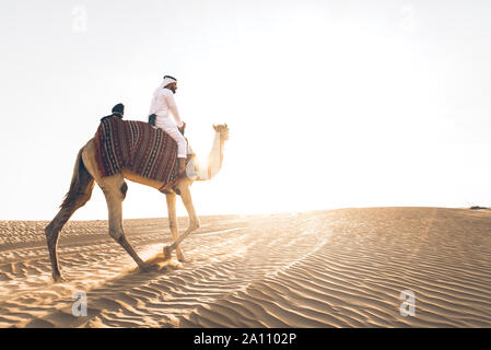 Uomo che indossa gli abiti tradizionali, tenendo un cammello sulla sabbia del deserto, a Dubai Foto Stock