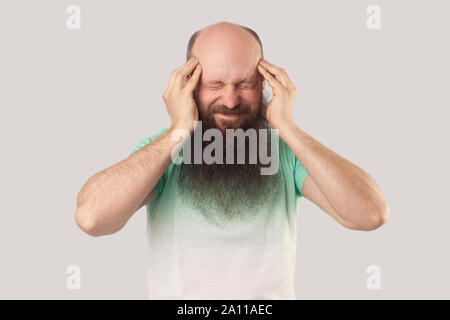 Mal di testa, pensando o confusione. Ritratto di mezza età uomo calvo con barba lunga in verde chiaro t-shirt e permanente tenendo la sua testa doloroso. interna Foto Stock