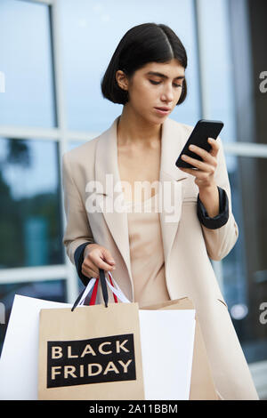 Vita ritratto di eleganti donna giovane azienda borse per lo shopping con il Black Friday e utilizza lo smartphone sul andare lasciando mall in stagione di vendita Foto Stock