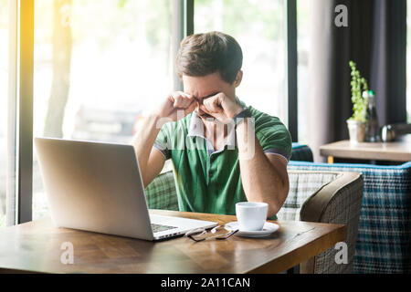 Giovane triste da soli proprietari in verde t-shirt seduta con il computer portatile, tenendo la testa verso il basso e strofinare i suoi occhi, o piangere. business e freelancing problema conc Foto Stock