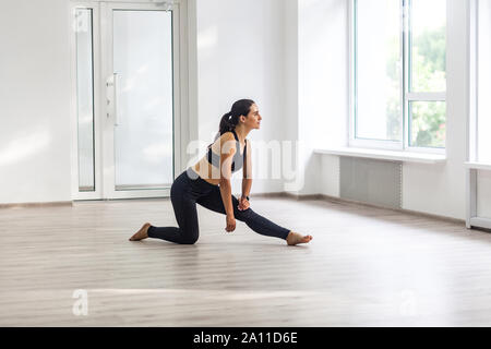 Giovane attraente sportivo da donna vestita di nero sportswear praticando sport esercizi, facendo balzi in avanti vicino alla finestra in palestra, stretching gambe e rilassante dopo Foto Stock