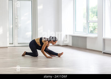 Giovane bella donna atletico di indossare abbigliamento sportivo nero praticando sport esercizi, facendo balzi in avanti vicino alla finestra in palestra, stretching gambe e rilassante dopo Foto Stock