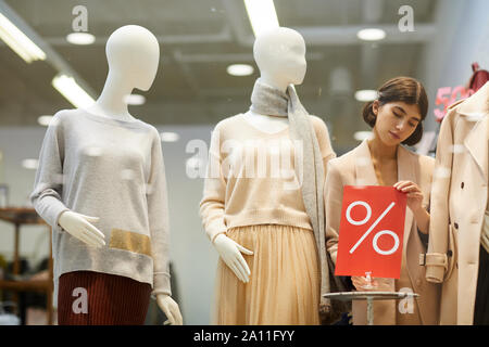 Ritratto di bella commessa di fissaggio vendita rosso segno e manichini vestito autunno Vesti durante l'impostazione window display in negozio, spazio di copia Foto Stock