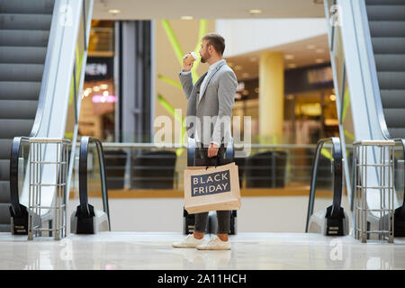 Vista laterale ritratto di moda uomo con le borse della spesa con il Black Friday iscrizione in piedi contro escalator in mall, spazio di copia Foto Stock