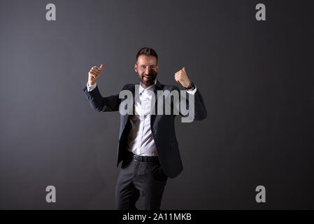 Un tifo bel giovane imprenditore in un vestito in piedi di fronte a uno sfondo grigio scuro in un studio. Foto Stock