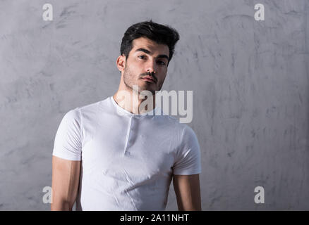 Un grave bel giovane in un bianco tshirt in piedi di fronte a un muro grigio in uno studio. Foto Stock