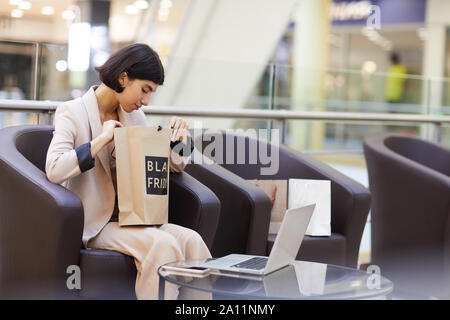 Vista laterale Ritratto di giovane e bella donna che guarda in sacchetto di carta e sorridente felicemente mentre godendo shopping spree in mall, spazio di copia Foto Stock