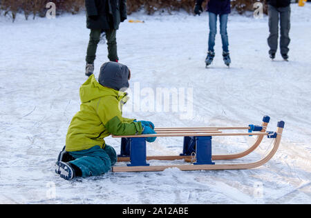 Piccolo Ragazzo seduto sul ghiaccio con sleigh guardando i pattinatori. Febbraio è il mese più freddo in tutto il Paesi Bassi Foto Stock