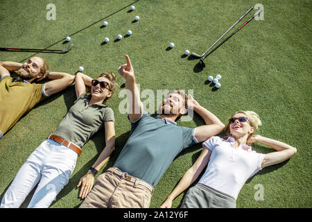 Il gruppo di un giovane e allegro amici giacente sul campo da golf con le palle e putters sull'erba, riposo e divertimento dopo il gioco Foto Stock