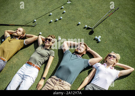 Il gruppo di un giovane e allegro amici giacente sul campo da golf con le palle e putters sull'erba, riposo e divertimento dopo il gioco Foto Stock