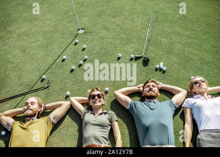 Il gruppo di un giovane e allegro amici giacente sul campo da golf con le palle e putters sull'erba, riposo e divertimento dopo il gioco Foto Stock