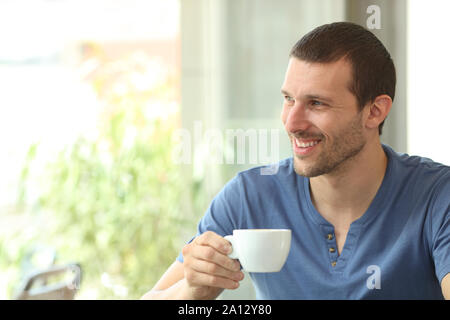 Uomo felice tiene una tazza di caffè guardando il lato attraverso una finestra in una caffetteria o home Foto Stock