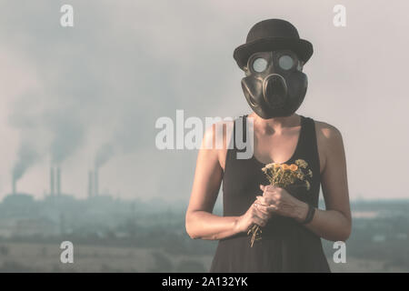 Giovane ragazza in un abito nero e una maschera a gas con fiori sullo sfondo del fumo ciminiere close up Foto Stock