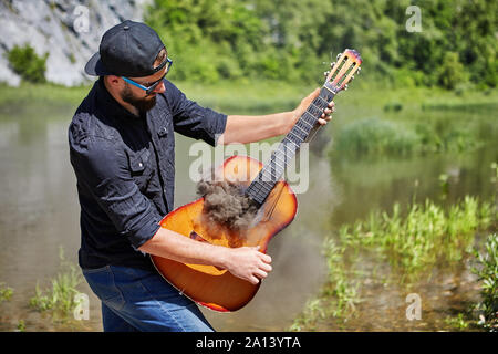 Un giovane uomo di 23 anni, indossando un cappello, occhiali da sole e un accartocciata camicia nera, riproduce una chitarra acustica da cui fumo spesso viene. Il chitarrista Foto Stock