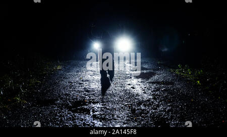 Un moody incappucciati figura di notte in ginocchio di fronte a dei fari delle automobili con la sua mano. Come heavy rain deriva verso il basso. Foto Stock