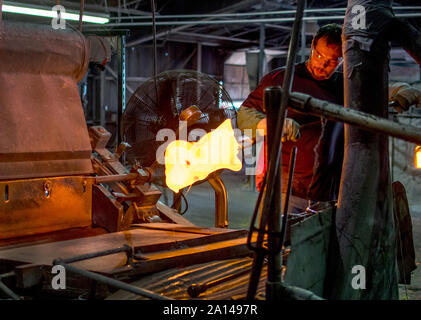 27 sett 2019 Kokomo Indiana America;un lavoratore di vetro utilizza strumenti di ferro e guanti pesanti per il sollevamento p un mucchio di vetro fuso. Egli ha poi impasti e volute i colori Foto Stock
