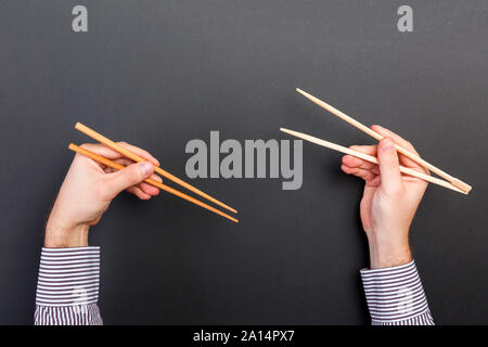 Immagine creativa delle bacchette di legno in due mani maschio su sfondo nero. Il giapponese e il cinese del cibo con copia spazio. Foto Stock