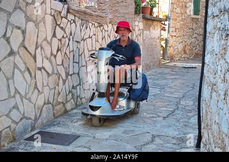 Ritratto di uomo anziano in sedia elettrica guida attraverso le strette strade di Primosten in Croazia Foto Stock