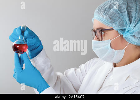 In un laboratorio di chimica, una femmina di farmacista sta analizzando un pallone con liquido rosso. Esperimento medico con un tubo. Farmacia lavorando su una prova con glov blu Foto Stock