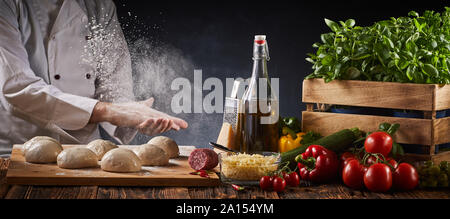 Lo Chef battendo la sua mani infarinata creando una nuvola di farina in aria come egli prepara le porzioni di pasta per pizza su una tabella con fresca ingredien assortiti Foto Stock