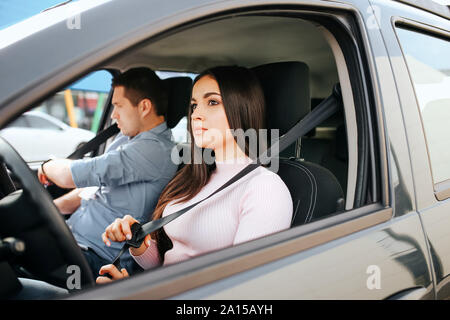 Maschio istruttore auto prende in esame giovane donna. Grave brunette tenere cintura in mano e guardare dritto. Guy preparare per la guida Foto Stock