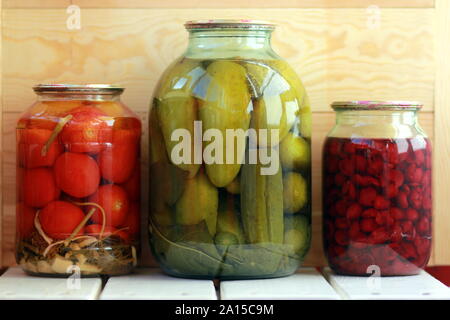 Tre vasi di cetrioli sottaceto, pomodori e confettura di ciliegie contro lo sfondo di legno Foto Stock