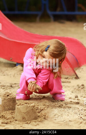 Un anno vecchio carina ragazza ricci in rosa tuta sportiva giocando e castelli di sabbia sul parco giochi per bambini Foto Stock