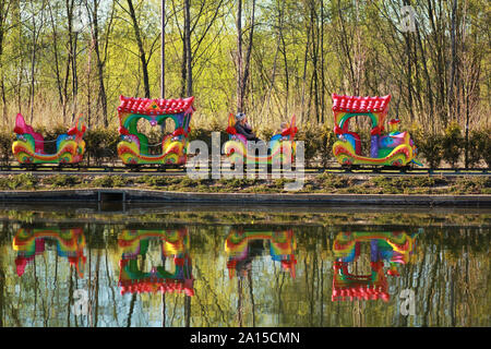 Madre e due bambini in sella a una coloratissima giostra treno nel Parco di primavera e la riflessione in un lago Foto Stock