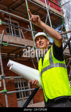 Builder maschio foreman, lavoratore geometra, ingegnere o architetto sul sito in costruzione tenendo i progetti di un edificio e puntamento Foto Stock