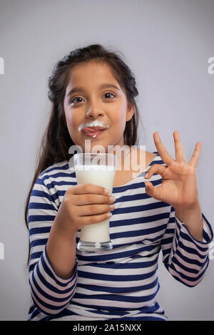 Ritratto di una ragazza sorridente con il latte di baffi tenendo un bicchiere di latte e facendo un segno OK con la mano Foto Stock