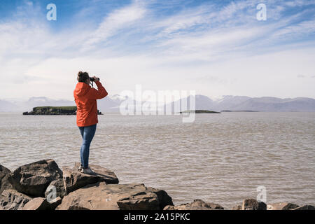 Giovane donna che lo standig massi, guardando attraverso il binocolo, Sud Est Islanda Foto Stock