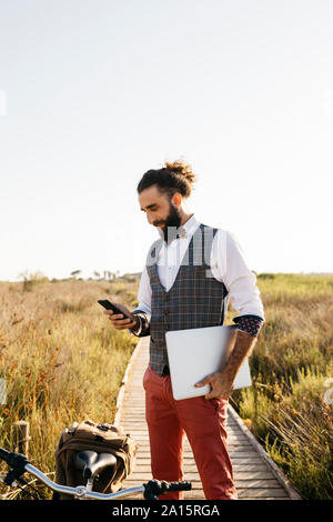 Ben vestito uomo in piedi su una passerella di legno in campagna accanto a una bicicletta tramite telefono cellulare Foto Stock