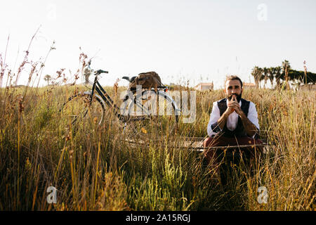 Ben vestito uomo seduto su una passerella di legno in campagna accanto a una bicicletta Foto Stock