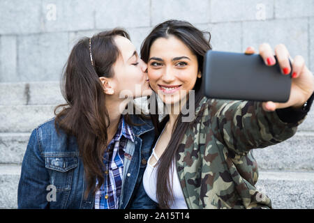 Donna baciando la guancia della sua amica mentre ella tenendo selfie Foto Stock