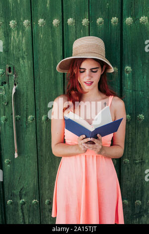 Ritratto di redheaded giovane donna in piedi di fronte a verde porta di legno la lettura di un libro in estate Foto Stock